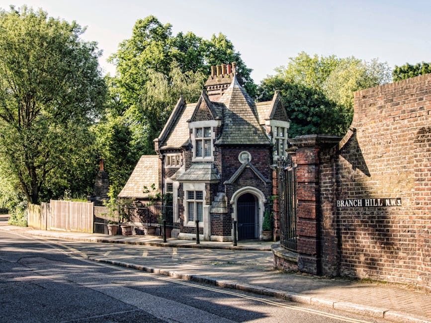 A narrow residential street scene showing a Victorian-style house with a steeply pitched roof, multiple chimneys, and ornate brickwork, situated behind a decorative wrought iron gate attached to a brick wall. The house is partially obscured by lush green trees and bushes lining the street, which has a kerbside pavement. There are no visible vehicles or moving equipment in the image. This setting exemplifies a typical home environment where removals by [COMPANY_NAME] may involve careful furniture transport, packing, and loading processes, especially in tight access properties like this. The street sign reads 'BRANCH HILL N.W.3', indicating the location, and the lighting suggests daytime with clear weather. The image captures a peaceful scene relevant to house removals and the logistical considerations associated with narrow access properties, as handled by [COMPANY_NAME] specializing in residential moving services.