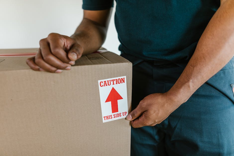 Close-up of a person dressed in dark clothing securing a cardboard box during a home relocation, with both hands gripping the box. The box has a red and white caution label indicating 'This Side Up' with an upward arrow. The setting appears to be inside a property, possibly near an entryway or hallway, with natural lighting illuminating the scene. The individual is in the process of packing or preparing the box for transportation as part of furniture transport or packing and moving activities, supported by the presence of other packed boxes and moving equipment in the background. This image exemplifies careful handling during loading and highlights safety measures taken during house removals, relevant to services offered by Removal Companies Streatham as they assist with efficient logistics and home relocation tasks near Streatham Common station.