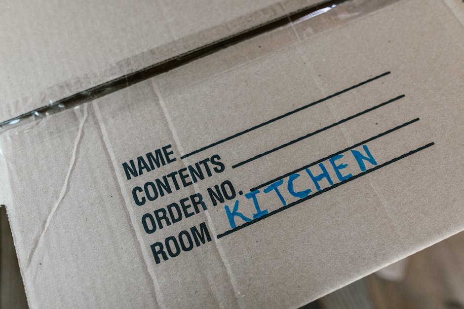 Close-up view of a cardboard moving box with printed labels and handwritten entries, including 'NAME', 'CONTENTS', 'ORDER NO.', and 'ROOM', with 'KITCHEN' written in blue marker next to the room label. The box appears to be part of a home relocation process, likely inside a property or on a pavement area, ready for loading onto a van managed by Removal Companies Streatham. The box is sealed with clear packing tape, and the background shows a glimpse of other boxes and packing materials, indicating an organized packing and moving operation. The scene emphasizes packing and furniture transport logistics involved in house removals or flat relocation, supporting the context of the webpage focused on SW16 removals tips for flats near Streatham Common station.