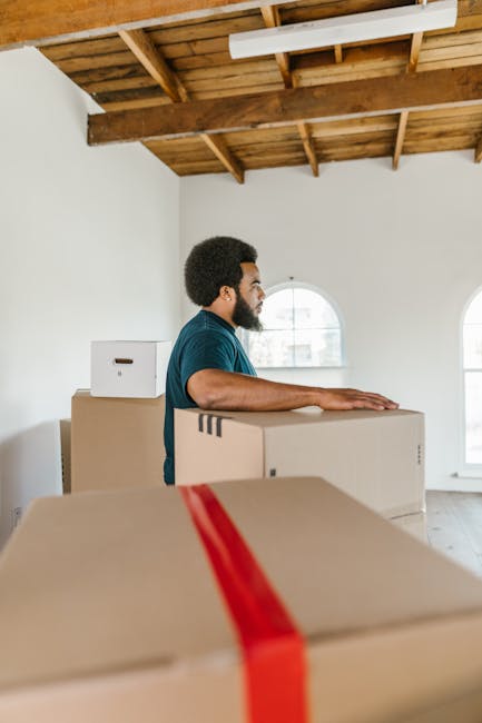 Close-up of a person dressed in dark clothing securing a cardboard box during a home relocation, with both hands gripping the box. The box has a red and white caution label indicating 'This Side Up' with an upward arrow. The setting appears to be inside a property, possibly near an entryway or hallway, with natural lighting illuminating the scene. The individual is in the process of packing or preparing the box for transportation as part of furniture transport or packing and moving activities, supported by the presence of other packed boxes and moving equipment in the background. This image exemplifies careful handling during loading and highlights safety measures taken during house removals, relevant to services offered by Removal Companies Streatham as they assist with efficient logistics and home relocation tasks near Streatham Common station.